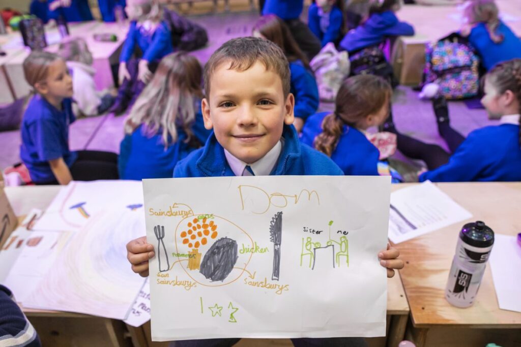 A school child holds up their drawing that was done as part of the activities during the From Field to Plate event. In the background other school children are visible interacting with one another.